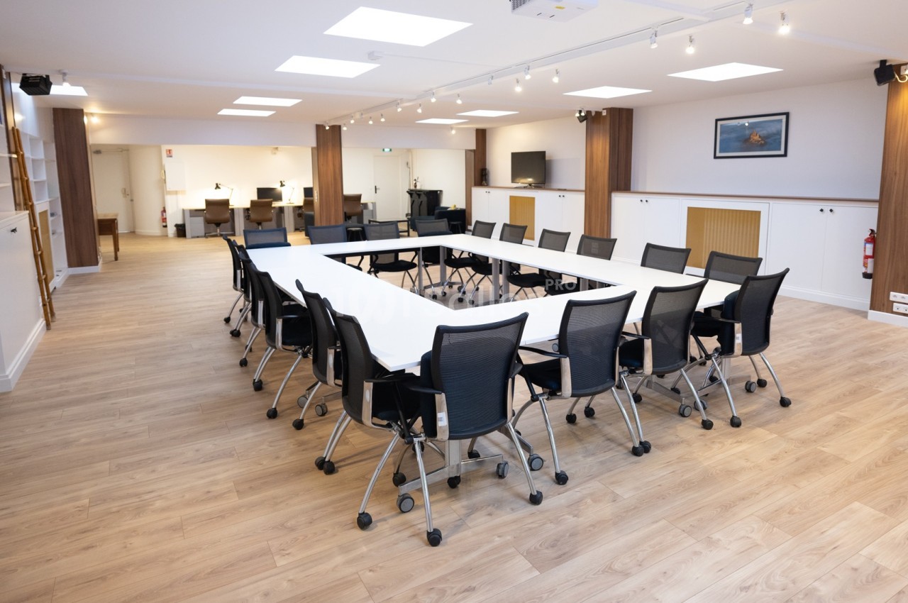 Salle de réunion lumineuse avec une grande table en U entourée de chaises noires sur un sol en bois clair.