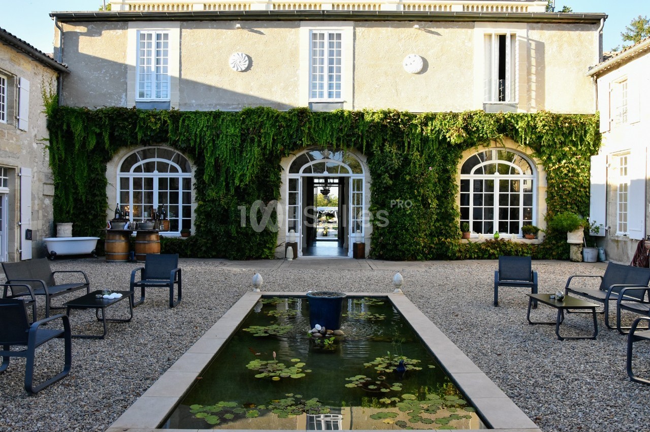 Façade d'une maison en pierre recouverte de lierre, avec une cour, un bassin et des chaises disposées autour.