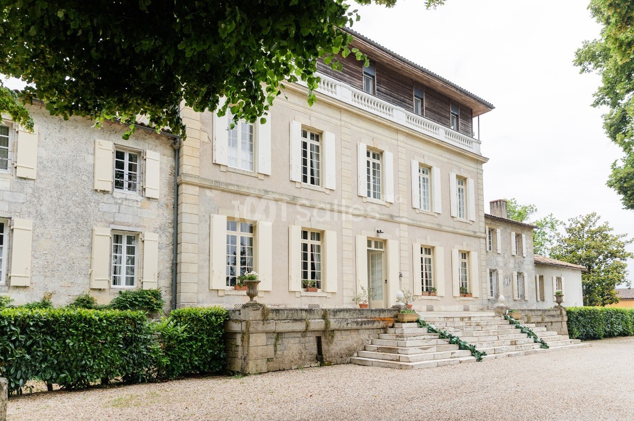 Façade d'une grande maison en pierre avec volets clairs, entourée de verdure et d'une allée gravillonnée.