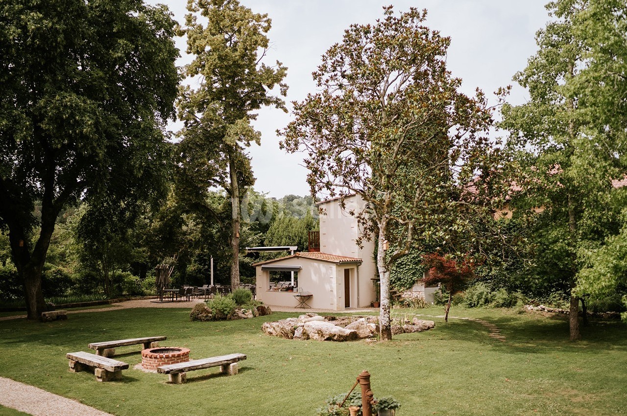 Jardin verdoyant avec bancs en bois, un foyer circulaire et une petite maison entourée d'arbres.