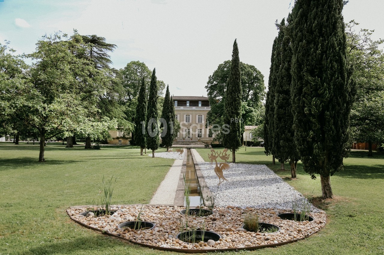 Allée bordée de cyprès menant à une grande maison, avec un bassin décoratif au premier plan dans un jardin verdoyant.