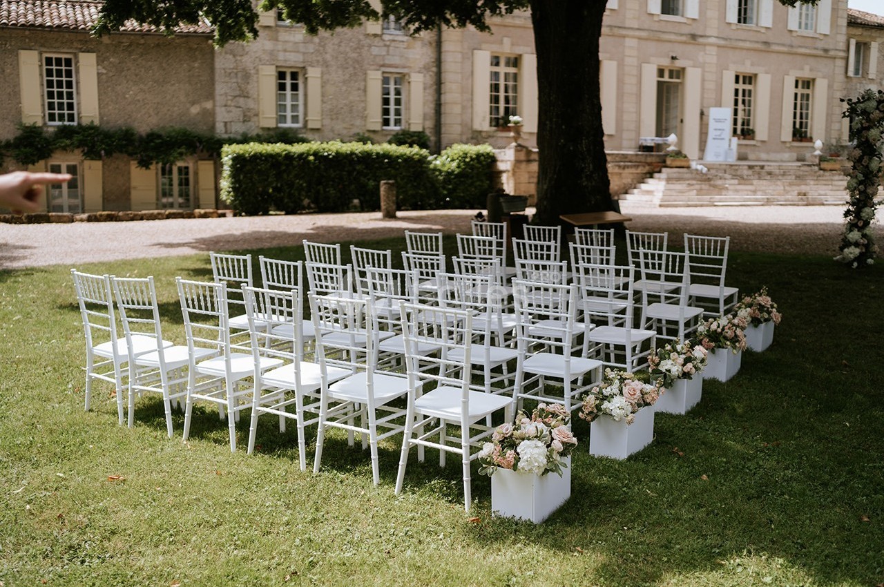 Chaises blanches disposées en rangées sur une pelouse, décorées de fleurs, devant un bâtiment en pierre.