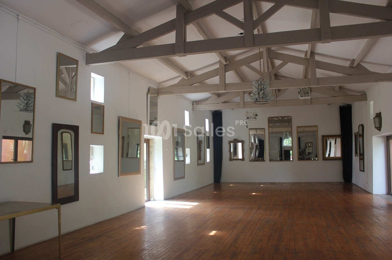 Salle lumineuse avec parquet en bois, murs blancs ornés de nombreux miroirs et poutres apparentes au plafond.