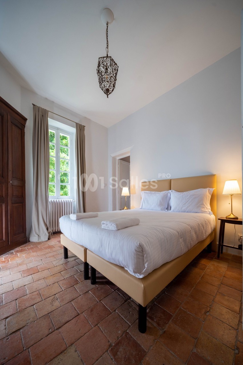 Chambre lumineuse avec lit double, sol en terre cuite, armoire en bois, lampes de chevet et fenêtre donnant sur un jardin.