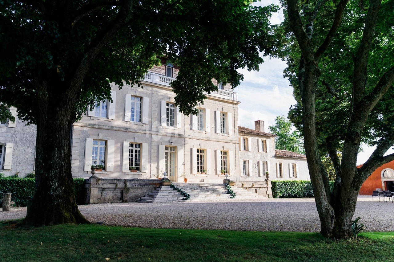 Façade d'une grande maison en pierre entourée d'arbres, avec une allée gravillonnée au premier plan.