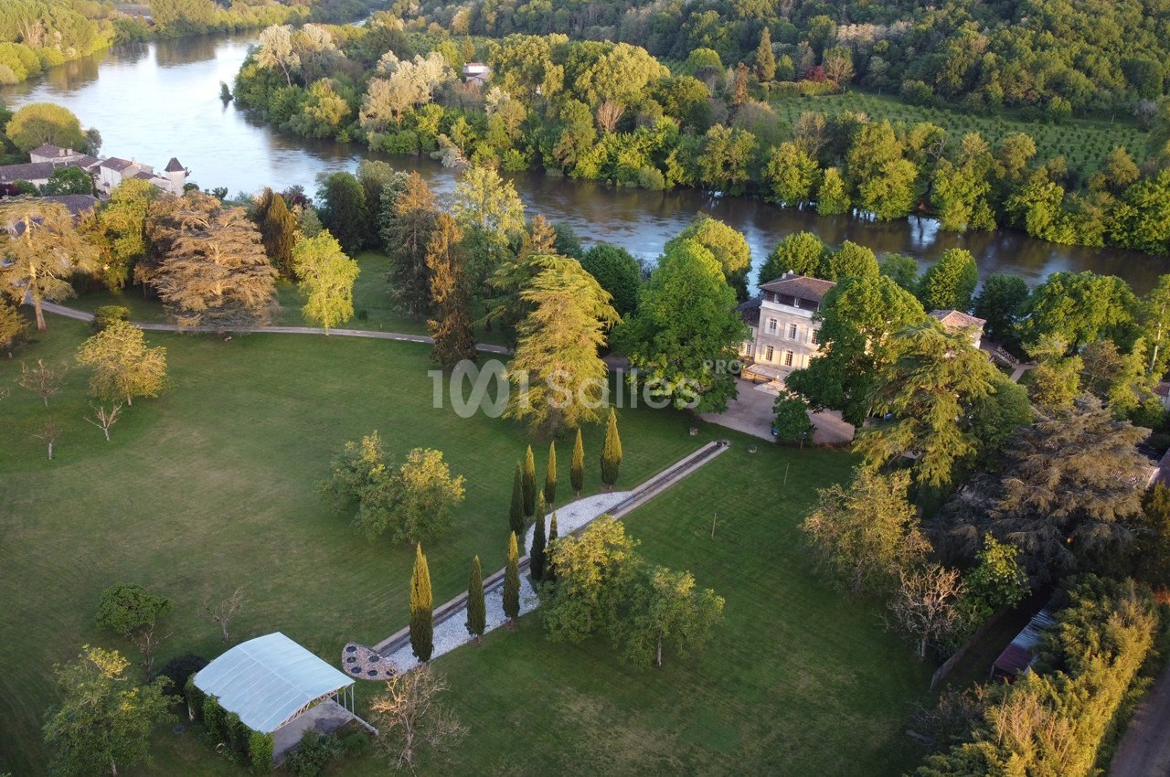 Vue aérienne d'un domaine avec une grande maison, un parc arboré, une rivière et une tente blanche dans un jardin.