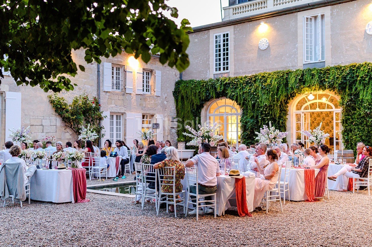 Invités assis à des tables décorées dans la cour d'une maison en pierre, lors d'une réception en plein air.