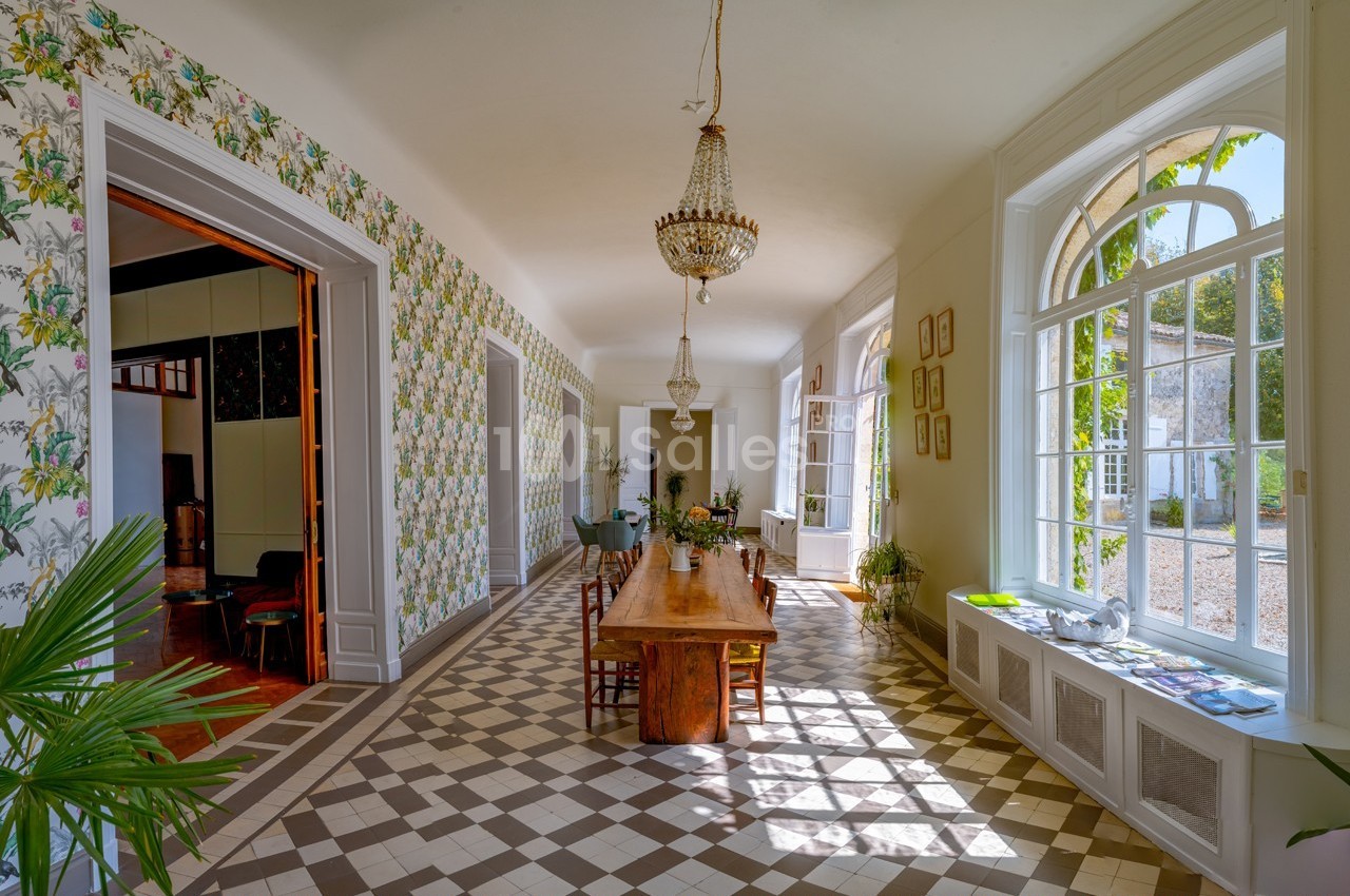 Salle lumineuse avec de grandes fenêtres, table en bois massif, sol en carreaux et murs ornés de papier peint floral.