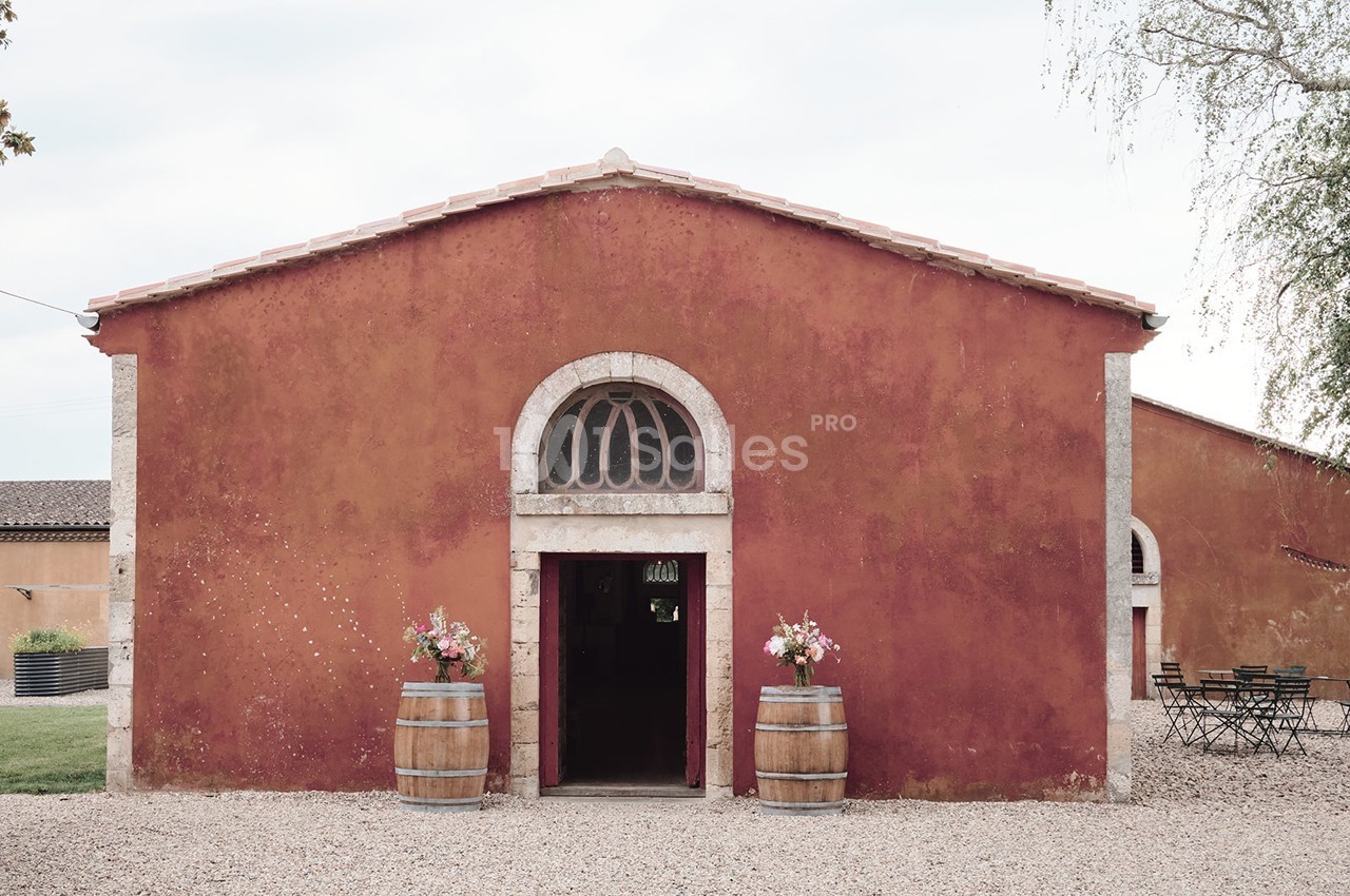 Façade d'un bâtiment en pierre rouge avec une porte voûtée, deux tonneaux décorés de fleurs et une cour gravillonnée.