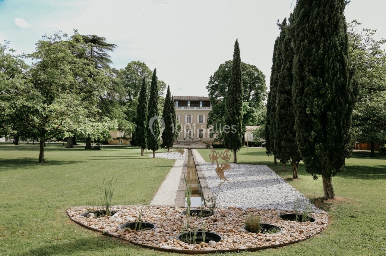 Allée bordée de cyprès menant à une grande maison entourée de pelouses et d'un jardin aménagé avec bassin et graviers.