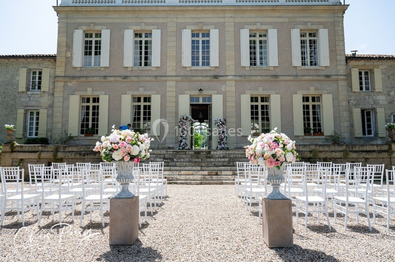 Allée de chaises blanches et décorations florales devant une grande bâtisse en pierre avec des fenêtres à volets.