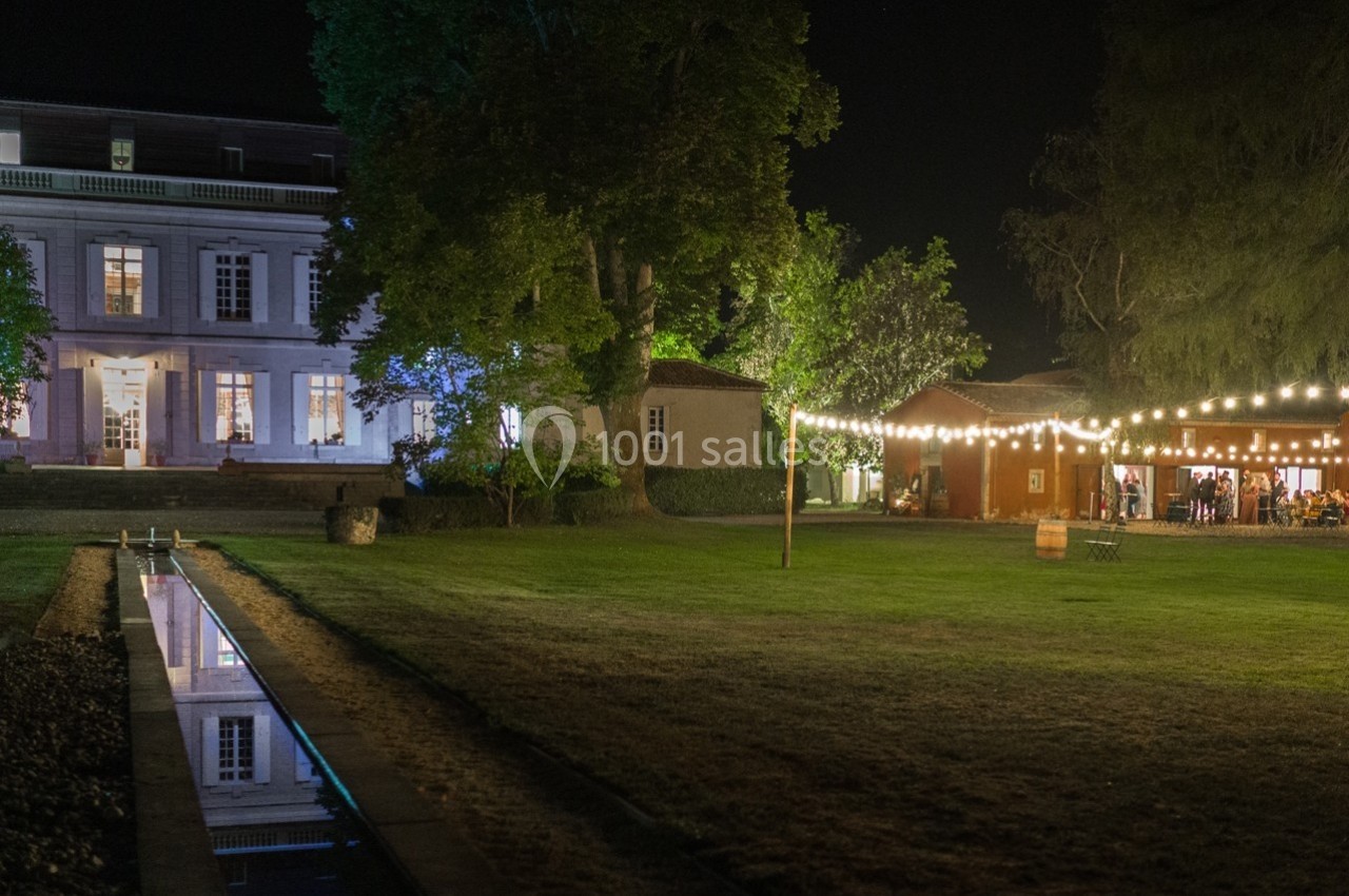 Grande maison éclairée et espace extérieur avec guirlandes lumineuses, pelouse et canal d'eau, de nuit.