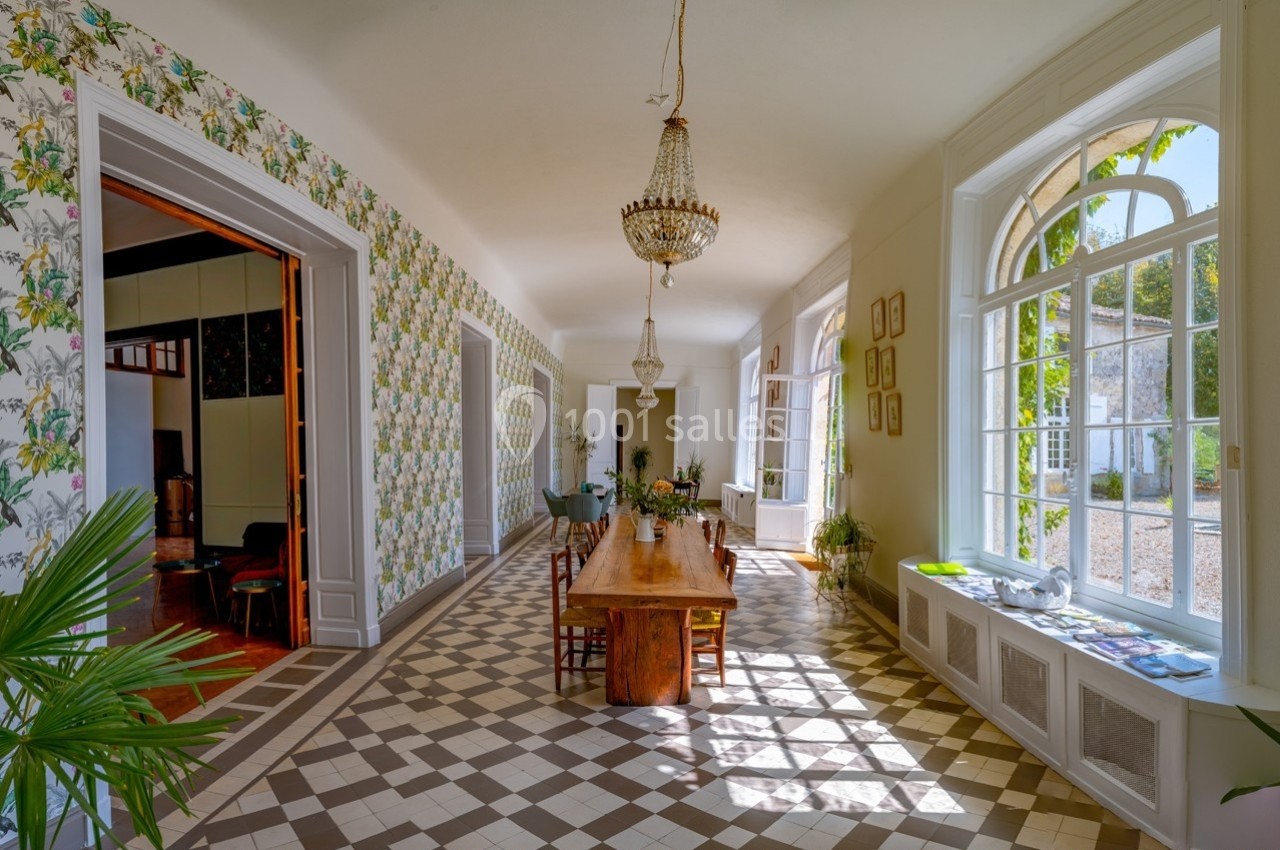 Salle lumineuse avec de grandes fenêtres, une table en bois, des plantes et un sol en carreaux géométriques.