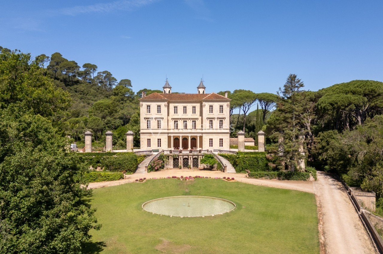 Vue d'un château entouré de jardins verdoyants avec une fontaine circulaire au centre, sous un ciel dégagé.