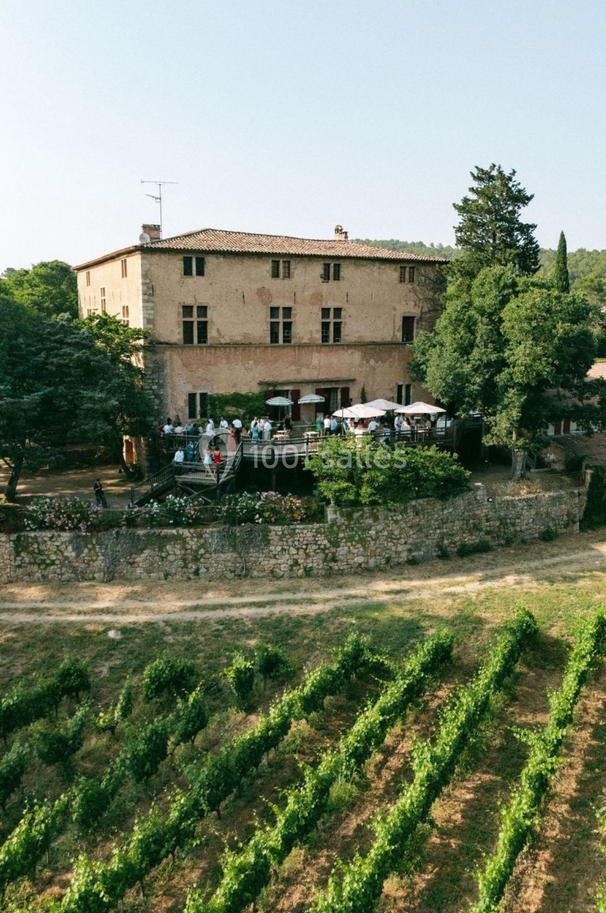 Vue aérienne d'un domaine avec une grande bâtisse en pierre, une terrasse animée et des vignobles au premier plan.