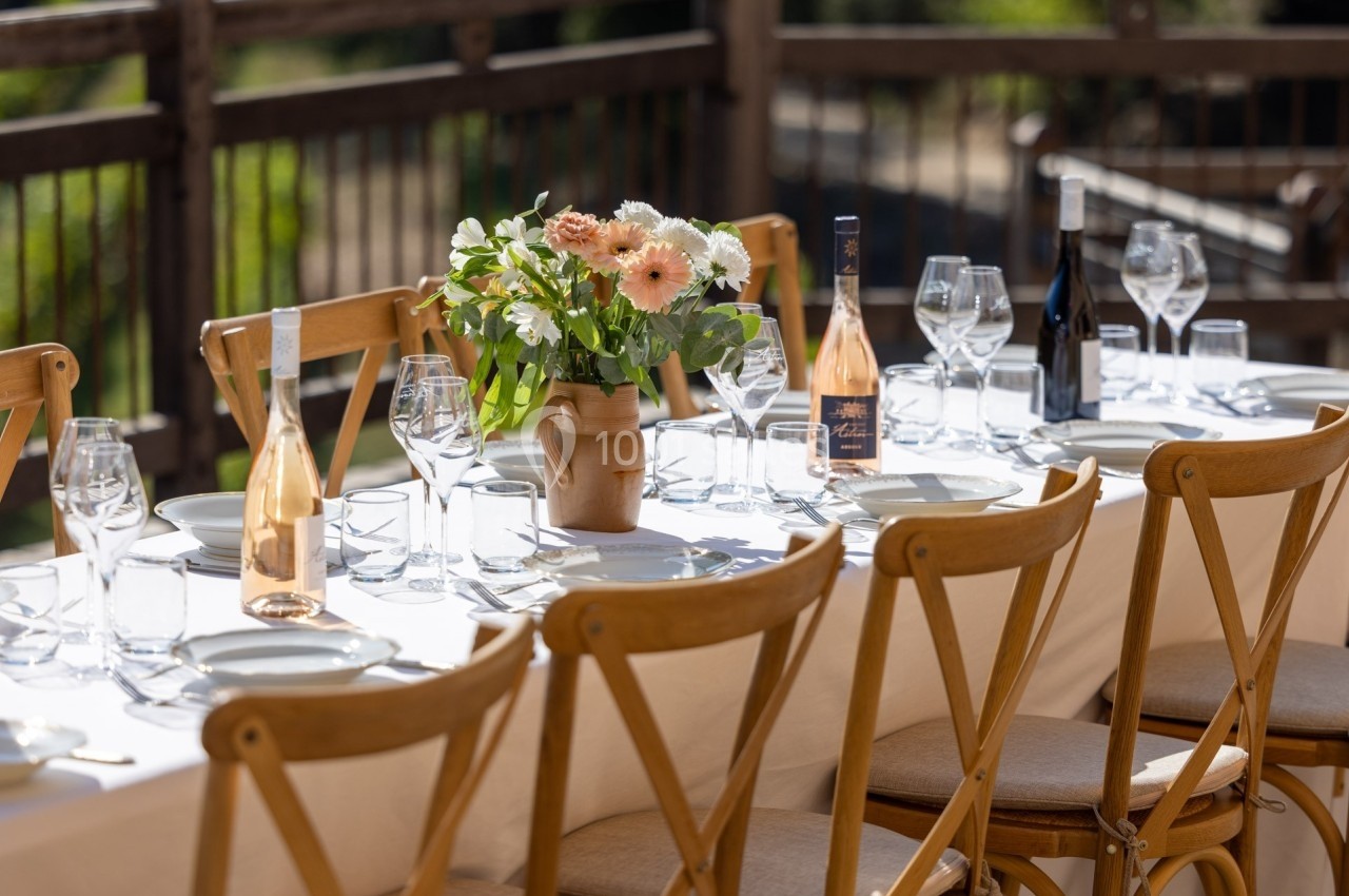 Table dressée en extérieur avec nappes blanches, chaises en bois, fleurs au centre et bouteilles de vin.