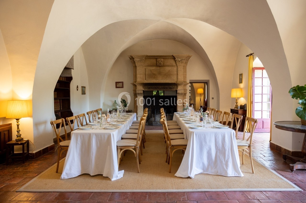 Salle voûtée avec deux longues tables dressées pour un repas, entourées de chaises en bois et éclairées par des lampes.