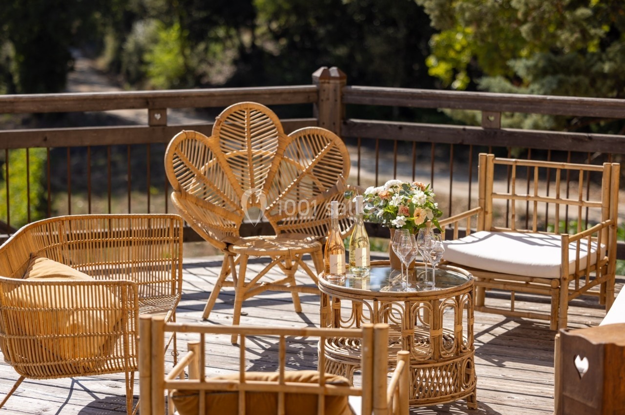Mobilier en rotin sur une terrasse en bois, avec table basse, fleurs, verres et bouteilles sous un environnement arboré.