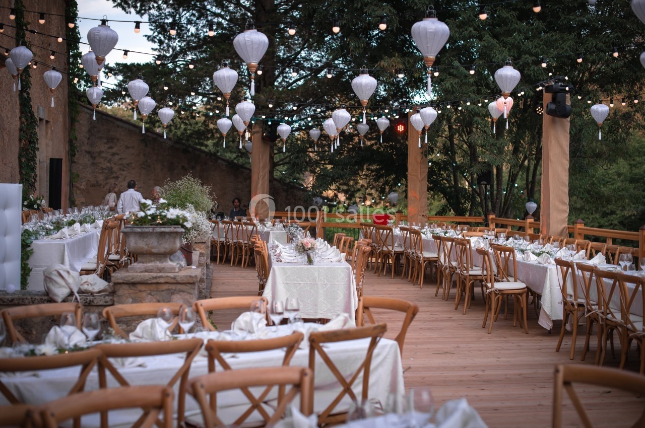 Terrasse décorée pour un événement avec tables dressées, chaises en bois et lanternes suspendues sous des arbres.