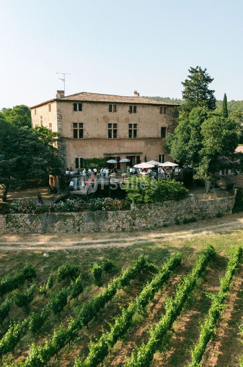 Vue aérienne d'un domaine avec un bâtiment en pierre, une terrasse animée et des vignes au premier plan.
