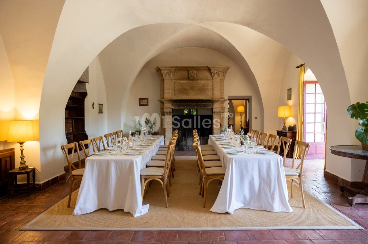 Salle voûtée avec deux longues tables dressées pour un repas, entourées de chaises en bois et éclairées par des lampes et la…