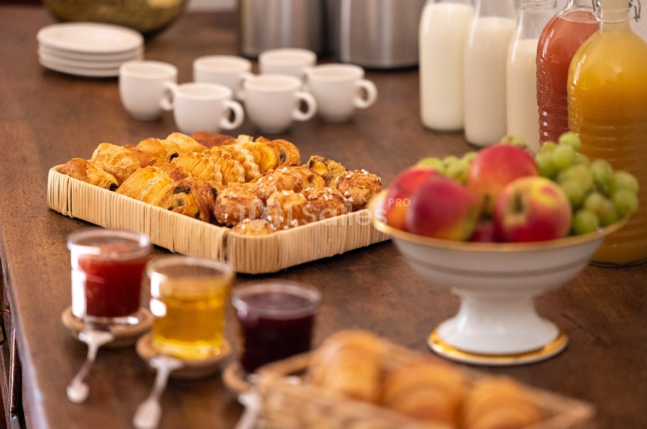 Plateau de viennoiseries entouré de confitures, fruits, jus et tasses sur une table en bois.