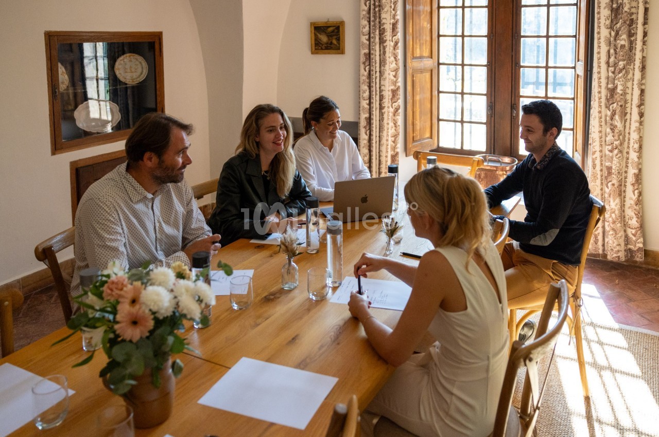 Un groupe de personnes discute autour d'une table en bois dans une pièce lumineuse avec des fenêtres et des fleurs.