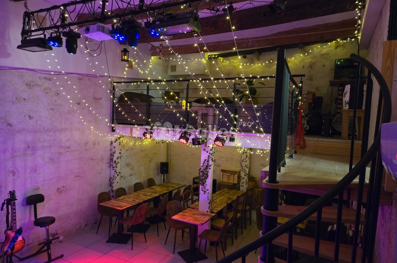 Salle avec tables en bois, guirlandes lumineuses suspendues, mezzanine et escalier en colimaçon.