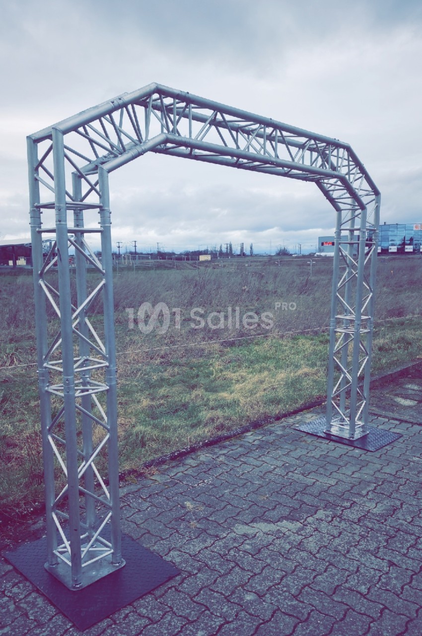 Structure métallique en forme d'arche installée sur un sol pavé, avec un paysage extérieur en arrière-plan.