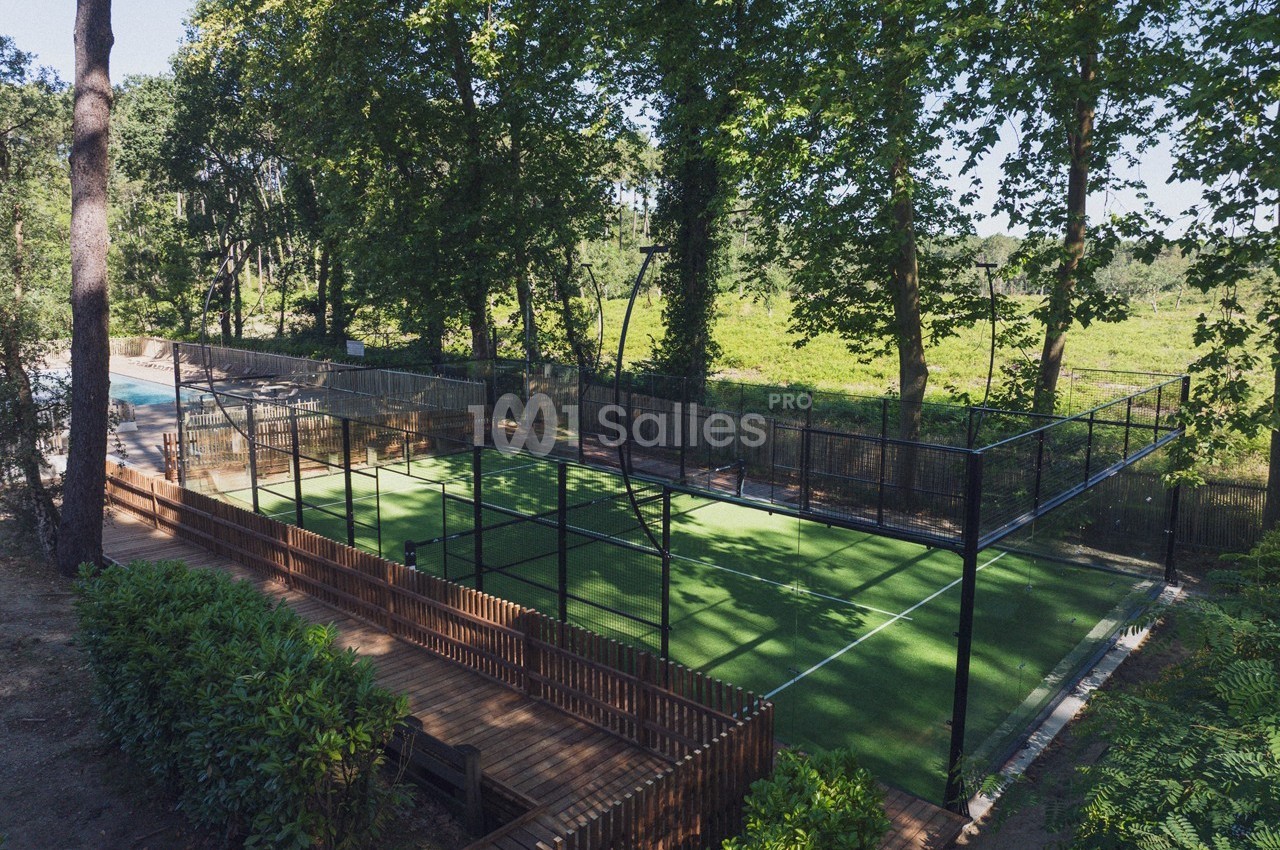 Terrain de padel clôturé entouré de végétation et d'arbres, avec un chemin en bois adjacent.