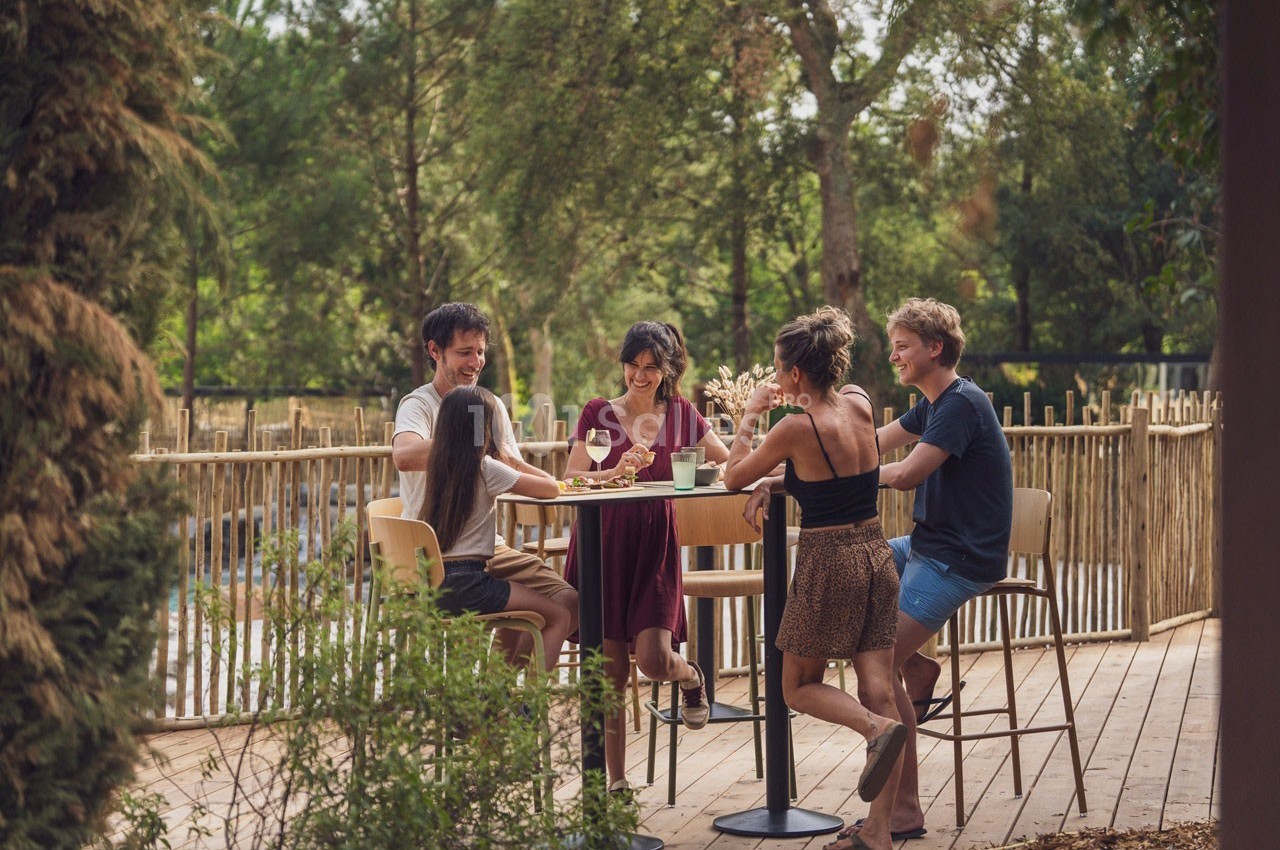 Des personnes discutent et partagent un repas autour d'une table haute en extérieur, entourées de verdure.