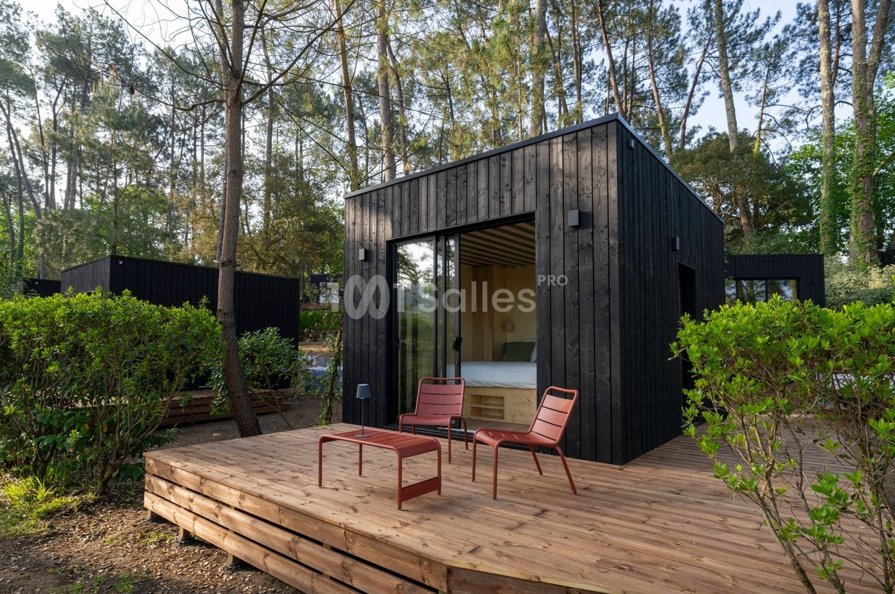 Cabane moderne en bois noir avec terrasse en bois, deux chaises rouges et table, entourée de végétation et de pins.