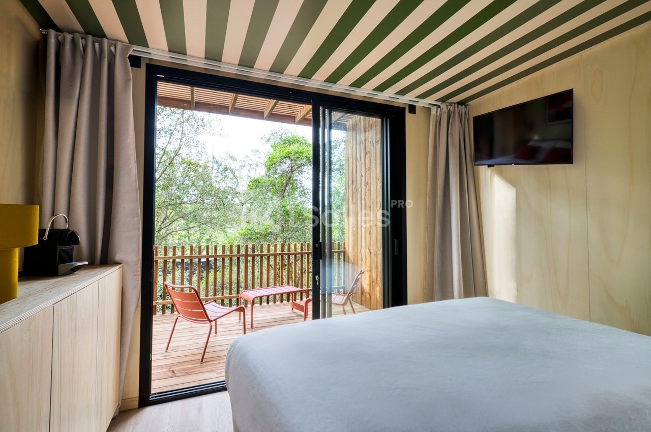 Chambre lumineuse avec lit, vue sur une terrasse en bois équipée de chaises, donnant sur un paysage arboré.