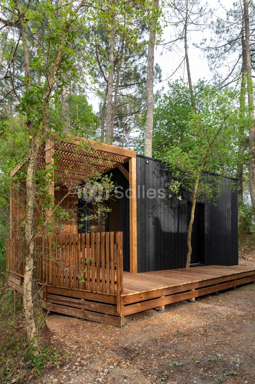 Cabane moderne en bois noir et naturel avec terrasse, entourée de pins et de végétation dense.