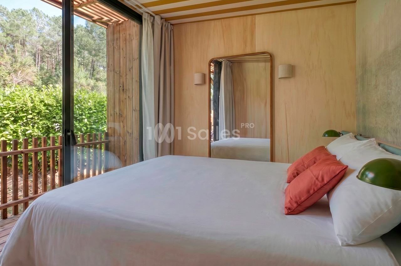 Chambre lumineuse avec lit double, miroir mural, grandes baies vitrées et vue sur une terrasse en bois et des arbres.