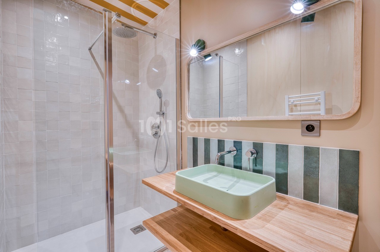 Salle de bain moderne avec douche vitrée, vasque verte sur un plan en bois et miroir rectangulaire.