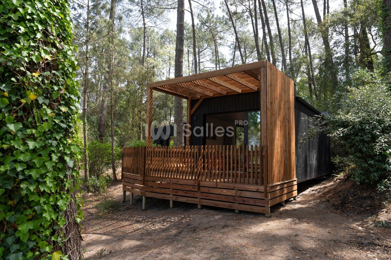 Cabane en bois moderne avec terrasse couverte, entourée de pins et de végétation dense en forêt.