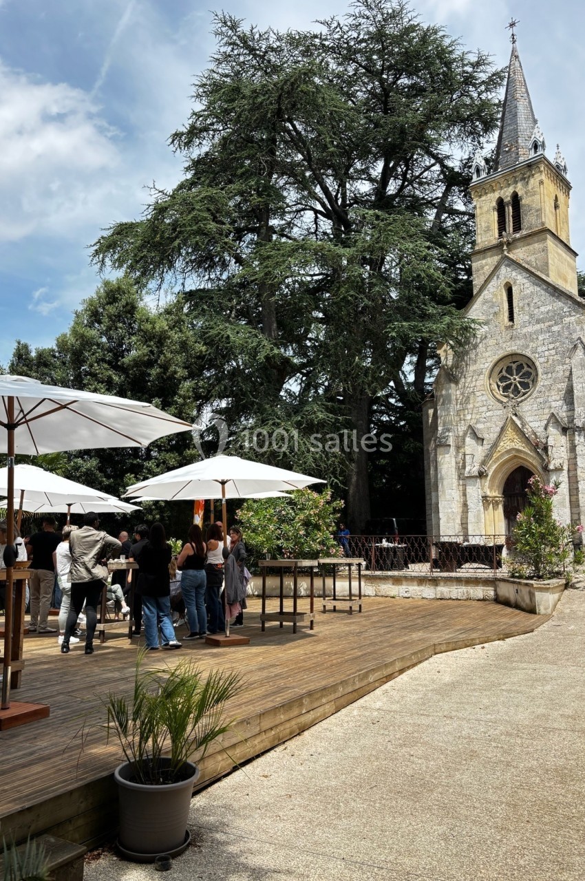 Terrasse en bois avec parasols et visiteurs près d'une petite église en pierre entourée d'arbres.