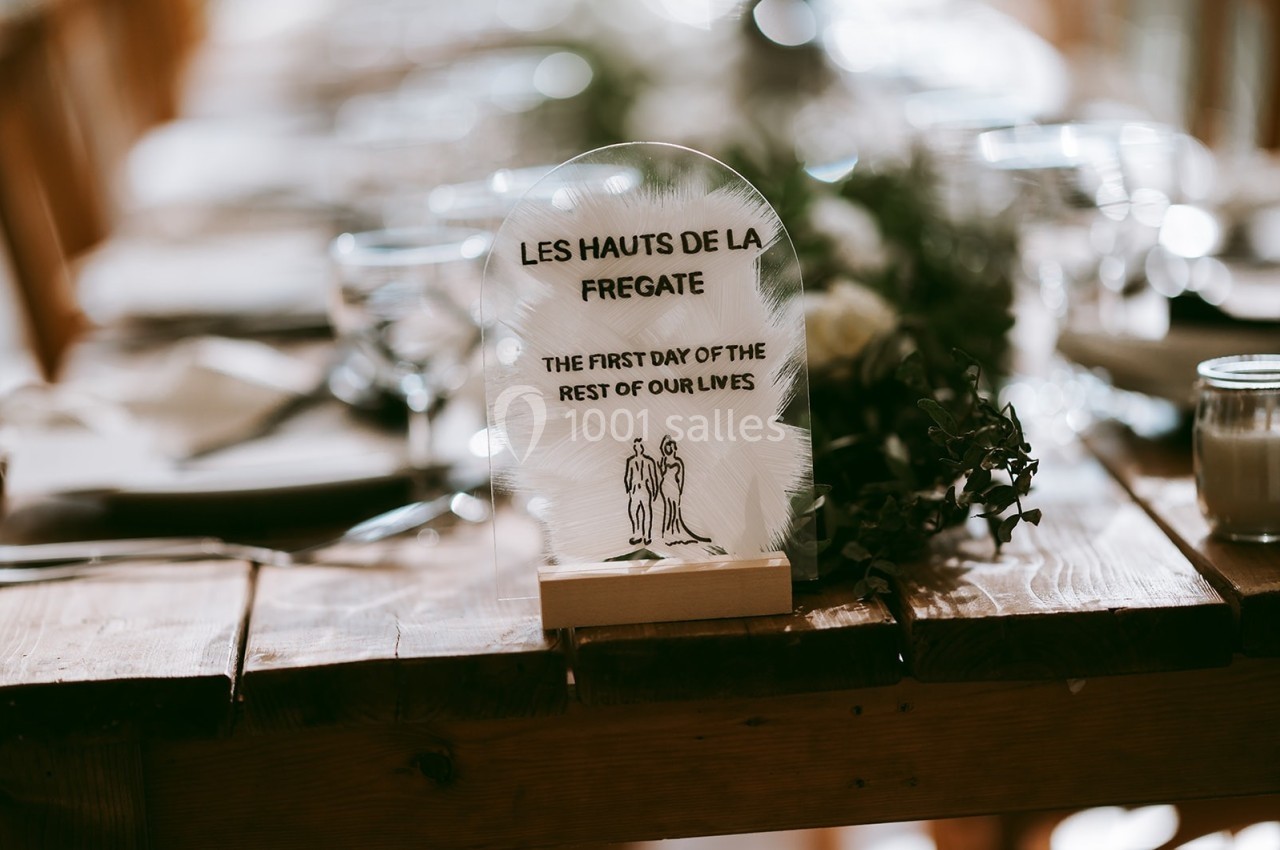 Carte de table en verre avec une inscription gravée, posée sur une table en bois décorée pour un événement.