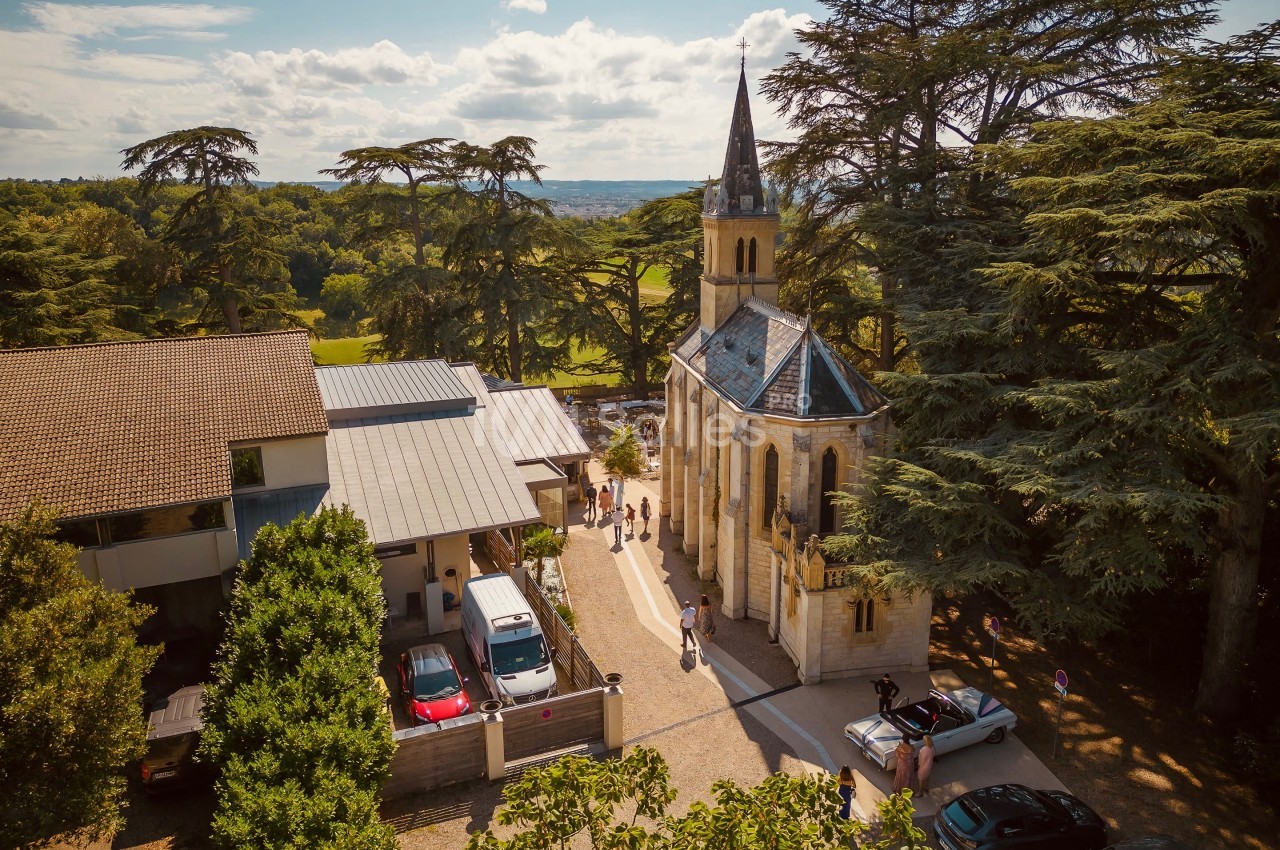 Vue aérienne d'une petite chapelle entourée d'arbres, avec des bâtiments modernes et des voitures à proximité.