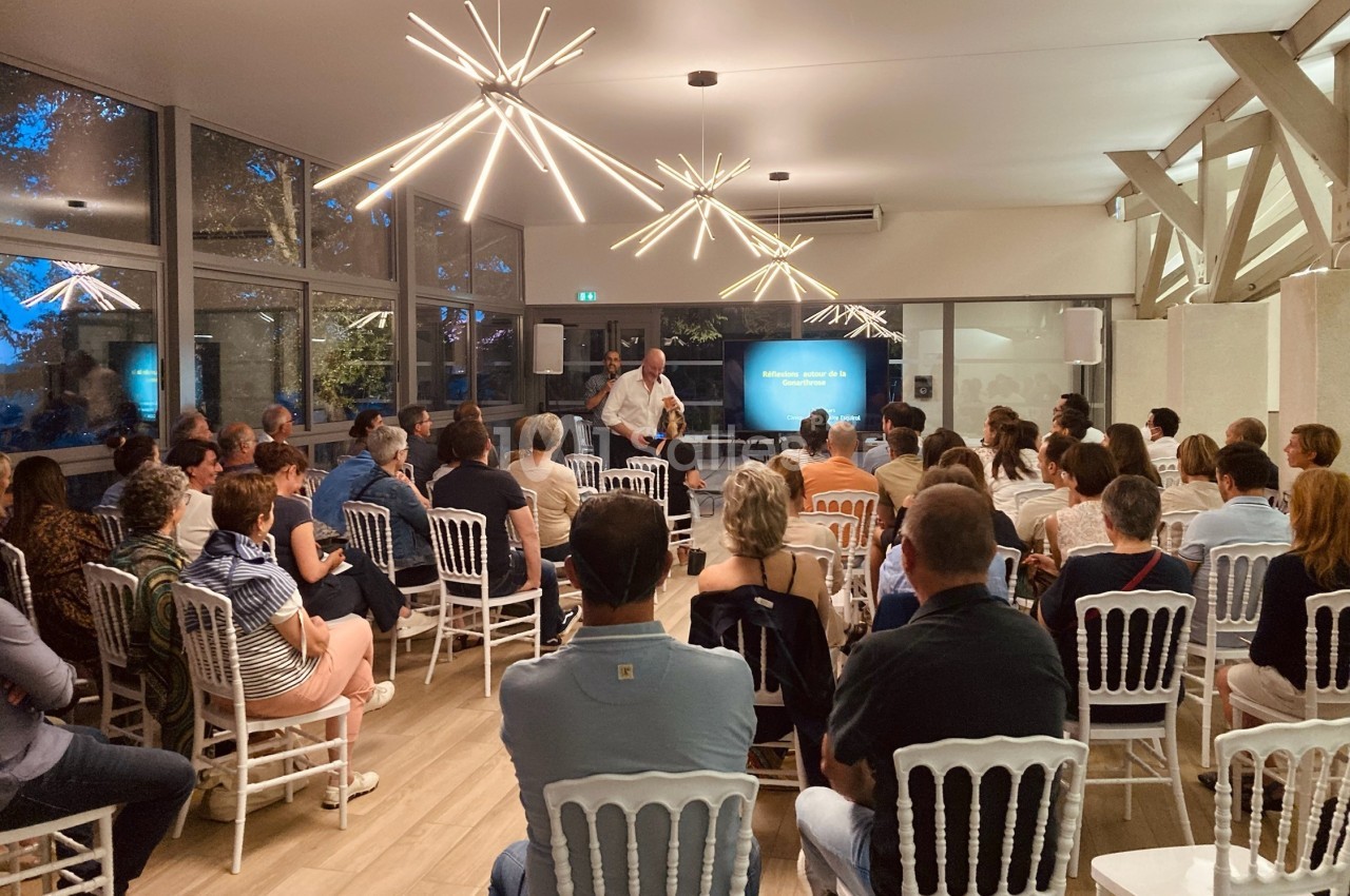 Un conférencier s'adresse à un public assis dans une salle lumineuse avec des chaises blanches et des éclairages modernes.