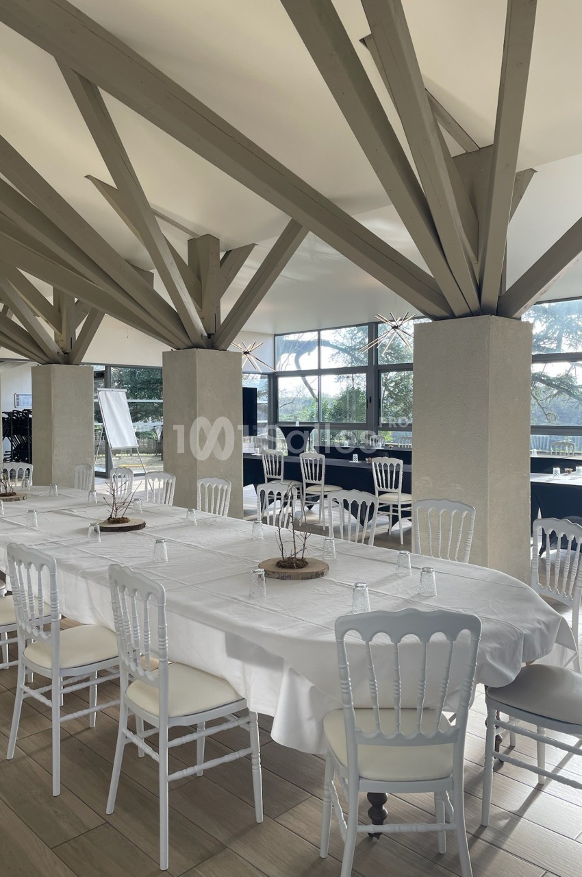 Salle lumineuse avec de grandes tables dressées de nappes blanches, entourées de chaises blanches et poutres apparentes.