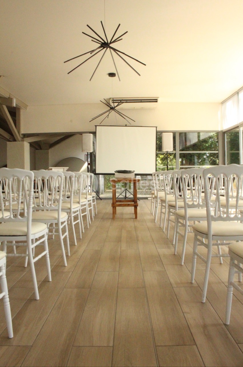 Salle lumineuse aménagée pour une présentation, avec des rangées de chaises blanches et un écran de projection.