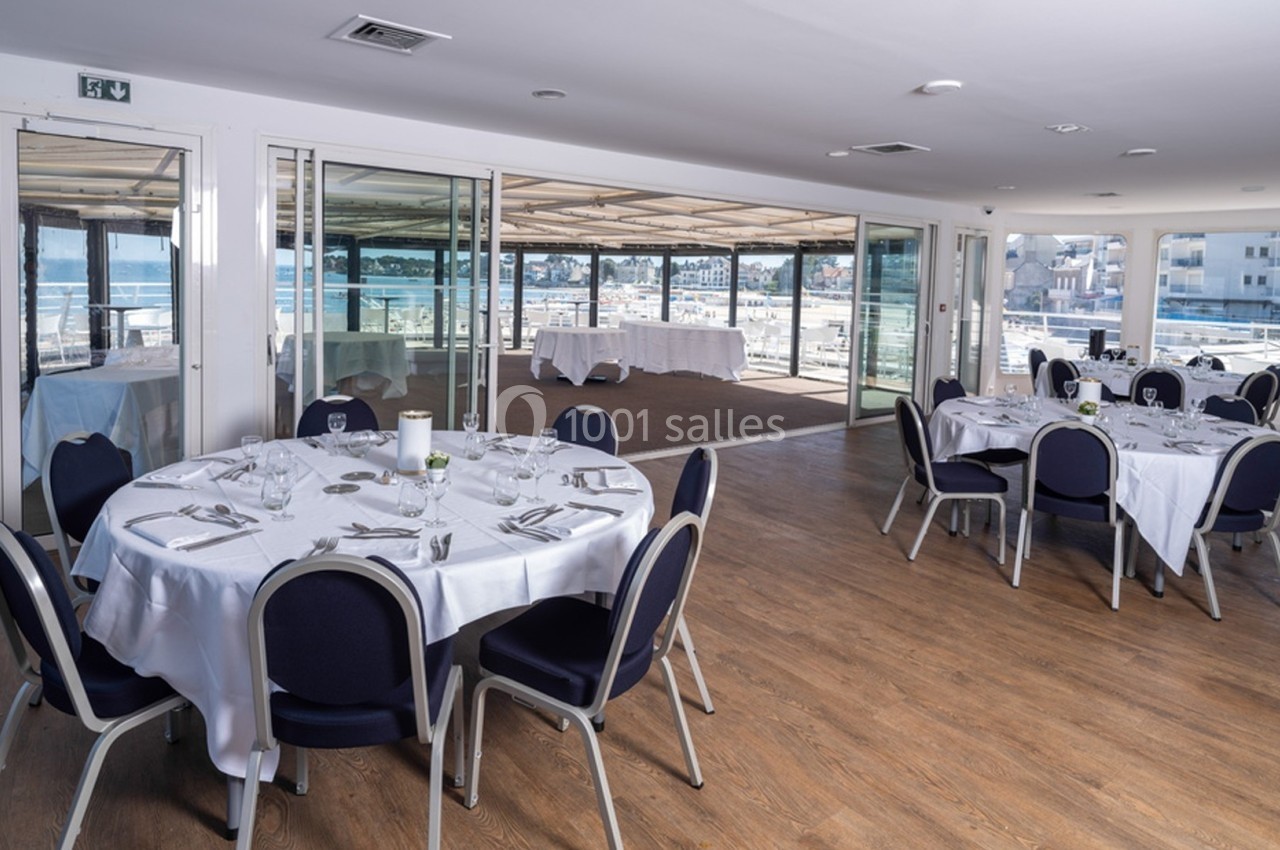 Salle de réception lumineuse avec tables rondes dressées, chaises bleues et vue sur une terrasse extérieure.