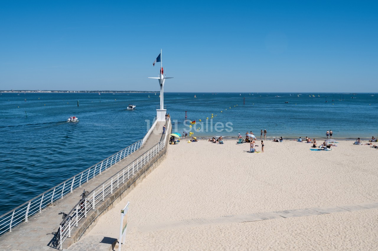 Digue avec croix blanche et drapeau, plage de sable avec baigneurs, mer turquoise et voiliers.