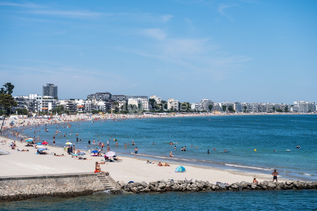 Plage du Pouliguen bondée avec baigneurs, sable blanc, digue en pierre et immeubles côtiers par temps ensoleillé.
