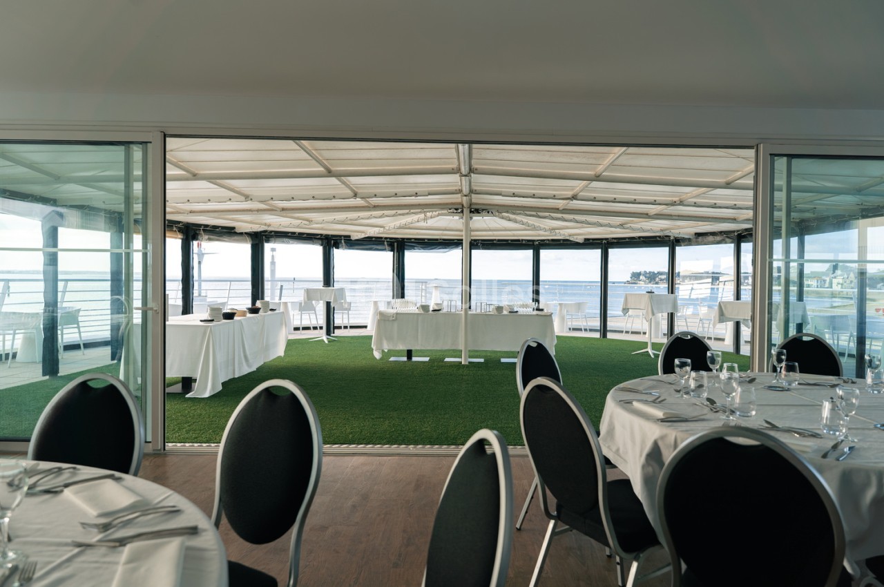 Salle de réception avec tables dressées, chaises noires, voilage rétractable et vue panoramique sur la mer.