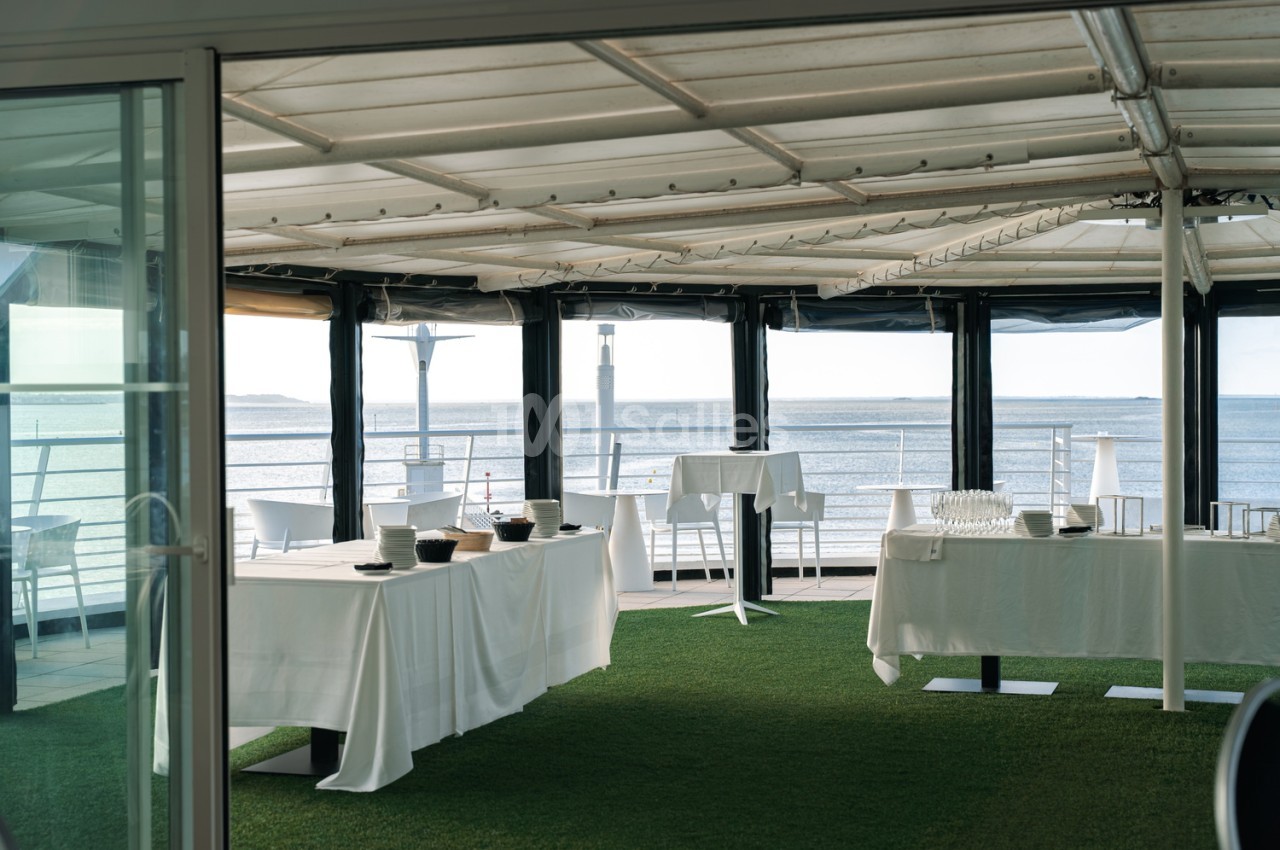 Salle aérée avec toiture rétractable, tables rondes dressées en blanc, chaises blanches et terrasse face à la mer.
