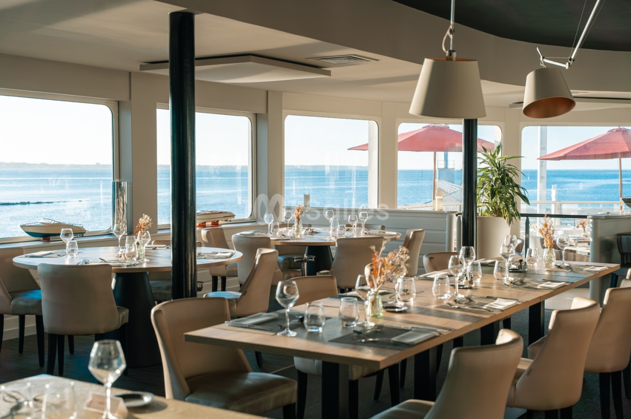 Salle de restaurant avec baies vitrées panoramiques sur la mer, tables dressées et chaises beiges, luminaires suspendus.