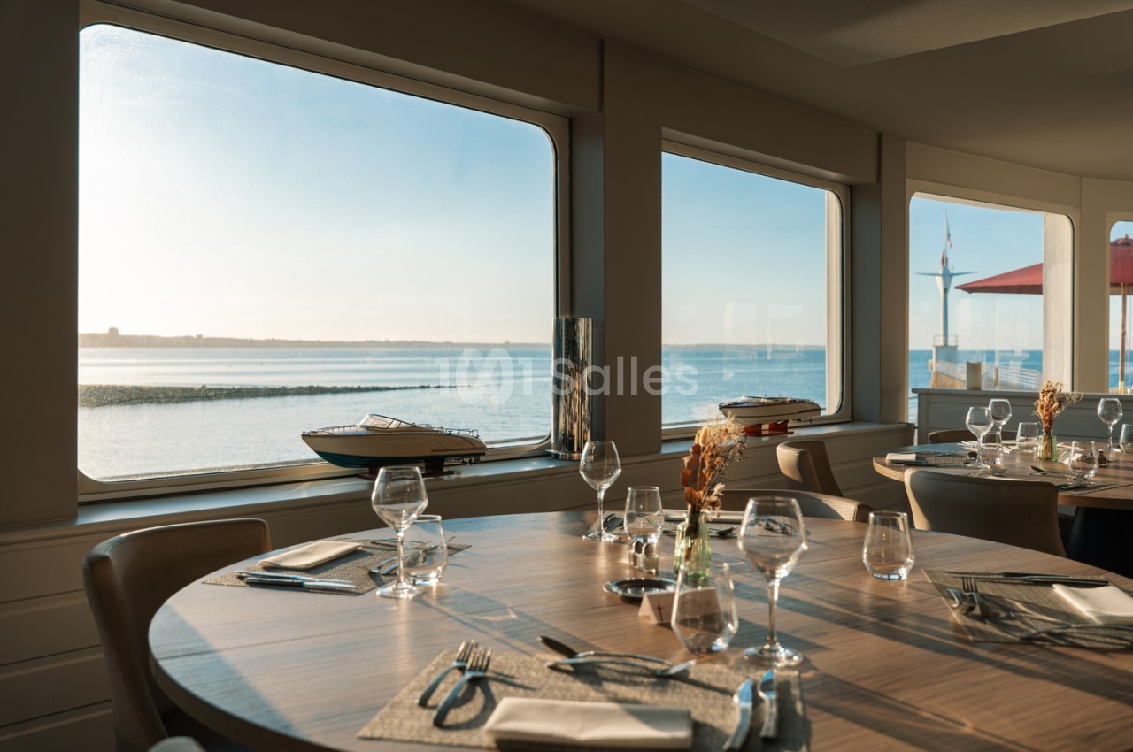 Salle de restaurant avec tables dressées en bois, vue panoramique sur la baie et bateaux de plaisance depuis les baies vitrées.
